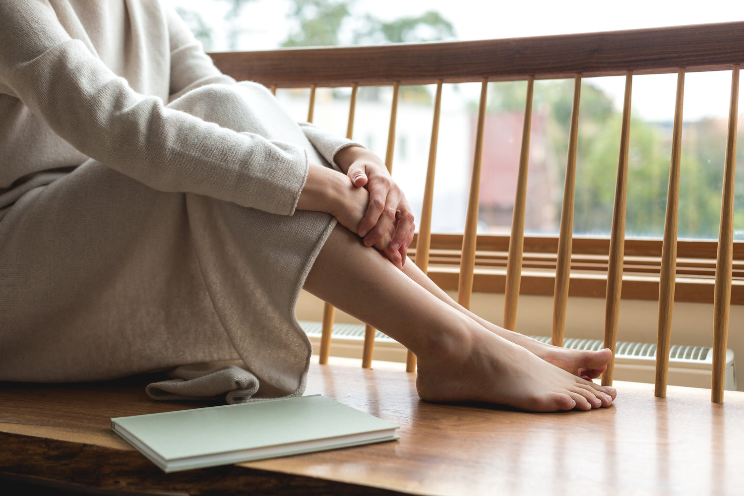 Woman sitting by a window holding her legs close, with a journal beside her in soft natural light
